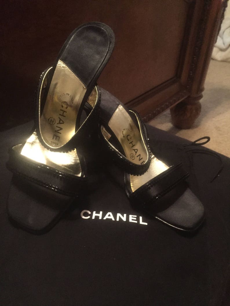 Pode incluir: Um par de sand&aacute;lias de salto alto de cetim preto com detalhes dourados e a palavra "CHANEL" na palmilha. As sand&aacute;lias est&atilde;o sobre uma bolsa de veludo preto com a palavra "CHANEL" impressa.