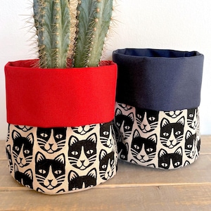 May include: Two fabric plant pots with a black and white cat face pattern. One pot has a red top and the other has a navy blue top.