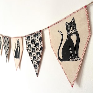 May include: A black and white cat bunting with a repeating pattern of cat faces and a single cat with a white chest sitting on a white background. The bunting is made of fabric and has red stitching.