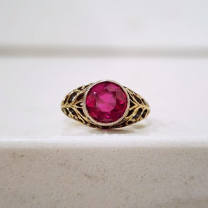 May include: A gold ring with a large, round, deep red ruby gemstone set in a filigree design.