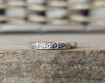 May include: A close-up of a gold-colored ring with a textured band. The ring is set on a wooden surface, with a blurred background. The ring's band has a hammered or textured design, adding visual interest to the piece.