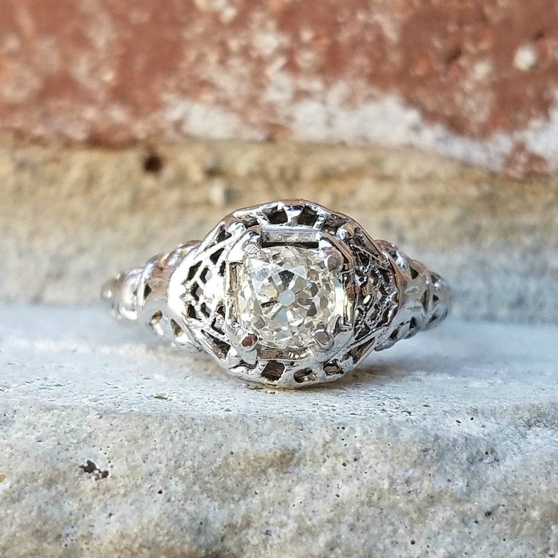May include: A vintage silver engagement ring featuring a large, clear, round diamond set in a filigree design.