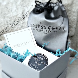 May include: A white box with a blue and white shredded paper filling. The box contains a small gray pouch with the text "Cypress Creek Antique Jewelry" and a small black and white sticker with the text "Cypress Creek Vintage" and a floral design.