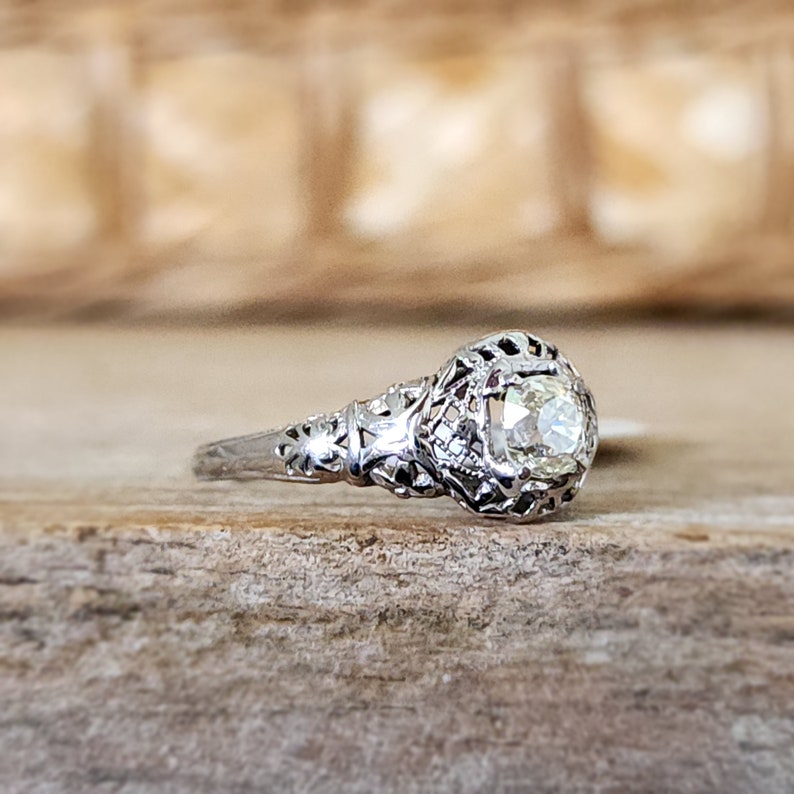 May include: A vintage engagement ring featuring a round brilliant cut diamond set in a filigree design. The ring is crafted from white gold and boasts a delicate, intricate design.