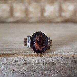 May include: A gold ring with a large, faceted, dark purple gemstone. The ring has a simple, elegant design.