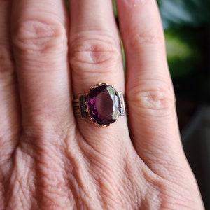May include: A gold ring with a large, oval-shaped purple gemstone set in a bezel setting. The ring has a decorative band with small details.