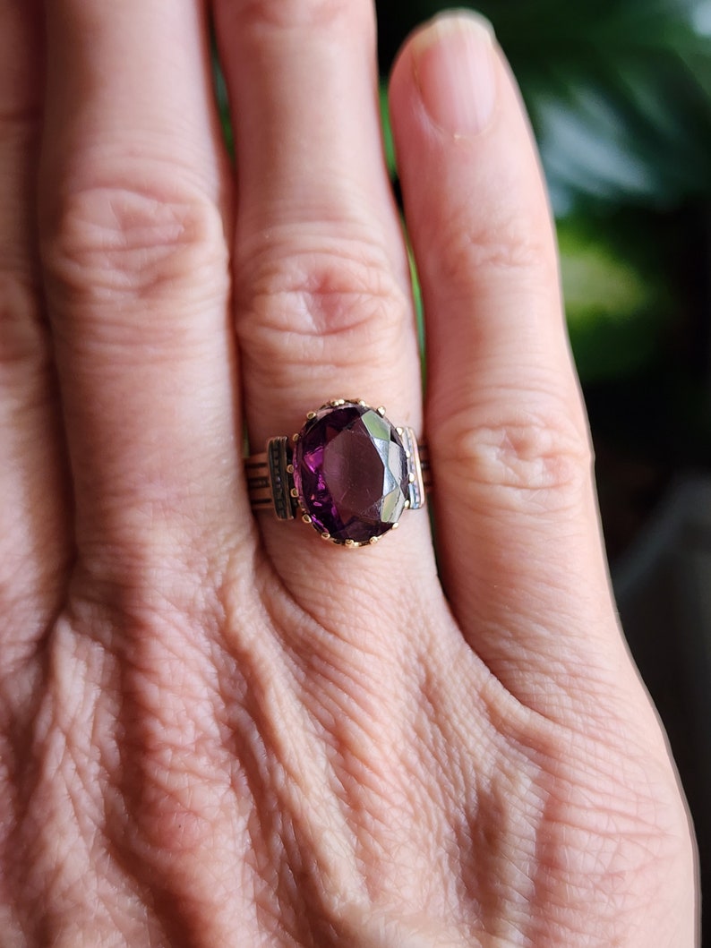 May include: A gold ring with a large, oval-shaped purple gemstone set in a bezel setting. The ring has a decorative band with small details.