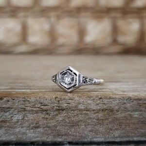 May include: A silver ring with a hexagonal setting featuring a small, clear diamond. The ring has a delicate design with small diamonds on the band.