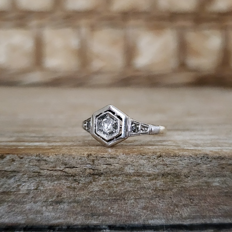 May include: A silver ring with a hexagonal setting featuring a small, clear diamond. The ring has a delicate design with small diamonds on the band.