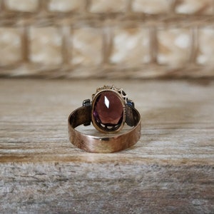 May include: A gold ring with a large, oval-shaped, purple gemstone set in a decorative, filigree setting. The ring is on a wooden surface.