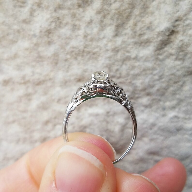 May include: A silver engagement ring with a delicate filigree design and a single round diamond.