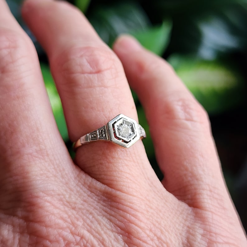 May include: A silver ring with a hexagonal setting featuring a small, round, clear diamond. The ring is set with smaller diamonds around the center stone.