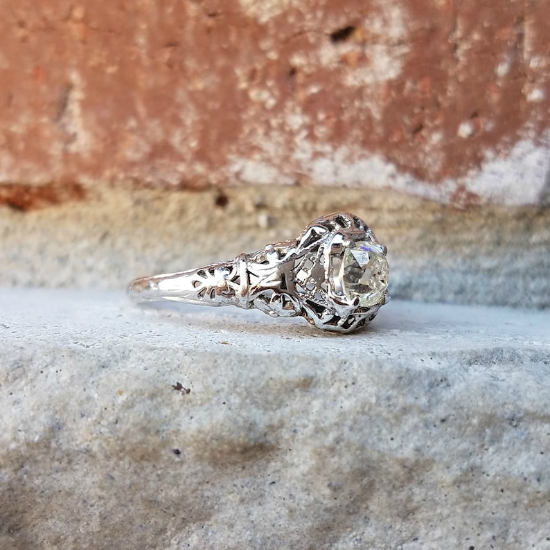 May include: A silver engagement ring with a delicate filigree design and a round, clear gemstone.