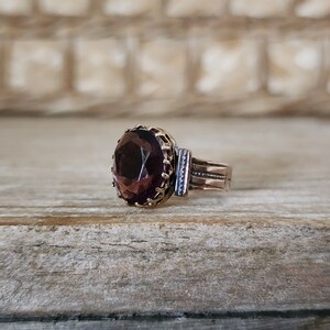 May include: A vintage ring with a dark purple gemstone set in a gold-toned bezel. The ring band is made of copper and has a delicate, textured design.