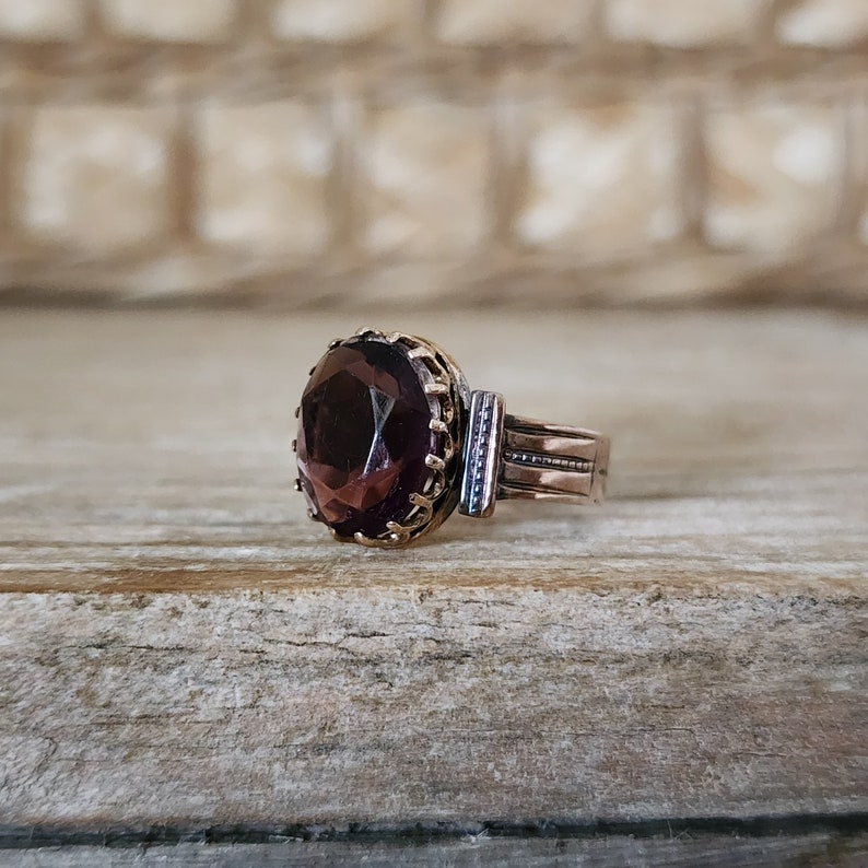 May include: A vintage ring with a dark purple gemstone set in a gold-toned bezel. The ring band is made of copper and has a delicate, textured design.
