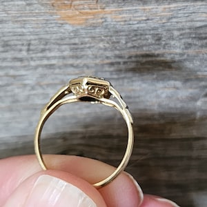 May include: A gold ring with a square-shaped stone set in a decorative setting. The ring has a delicate design with intricate details.