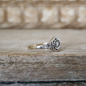 May include: A delicate gold ring with a hexagonal setting featuring a small, clear diamond surrounded by smaller diamonds. The ring is set against a rustic wooden background.