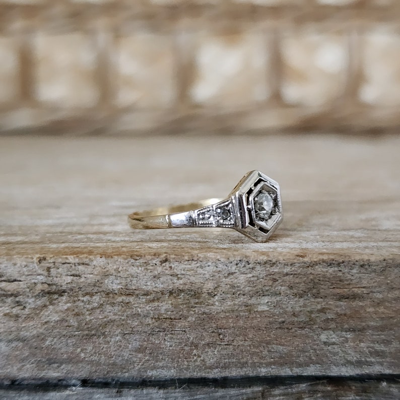 May include: A delicate gold ring with a hexagonal setting featuring a small, clear diamond surrounded by smaller diamonds. The ring is set against a rustic wooden background.