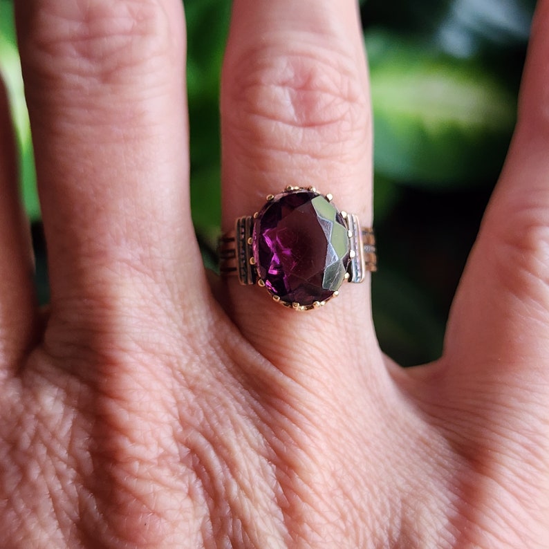 May include: A gold ring with a large, oval-shaped, purple gemstone set in a bezel setting. The ring has a decorative band with a gold and silver design.