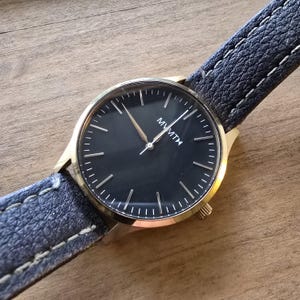 May include: A gold-coloured wristwatch with a black face and a navy blue leather strap. The watch face displays the brand name "MVMT" in white. The strap is stitched with white thread.