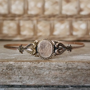 May include: A gold-toned bracelet with a circular, blank, metal locket in the center. The locket is surrounded by ornate, detailed designs on either side.
