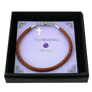 May include: A brown braided leather bracelet with a silver-toned cross charm and metal clasp, presented on a light purple card inside a black box. The card features the text To a Special Boy and With Love, along with a small purple heart icon.