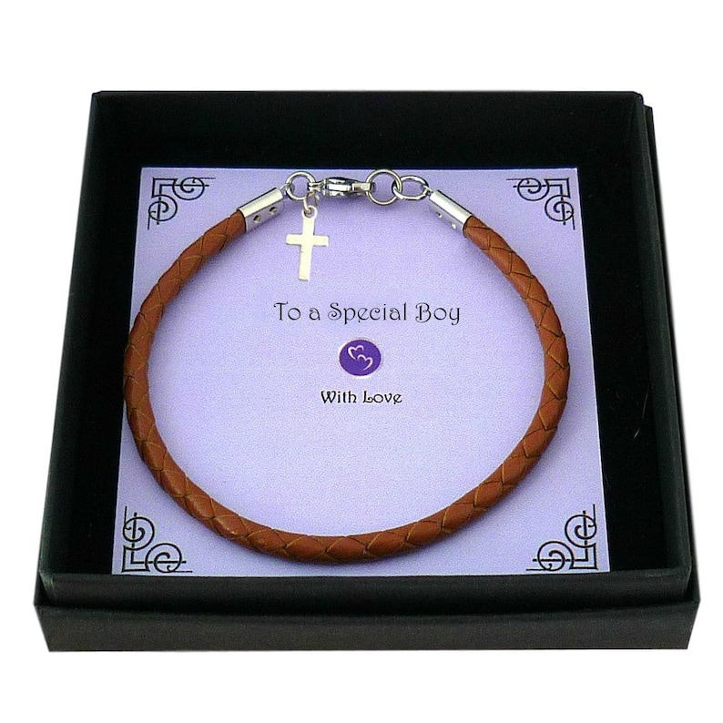 May include: A brown braided leather bracelet with a silver-toned cross charm and metal clasp, presented on a light purple card inside a black box. The card features the text To a Special Boy and With Love, along with a small purple heart icon.