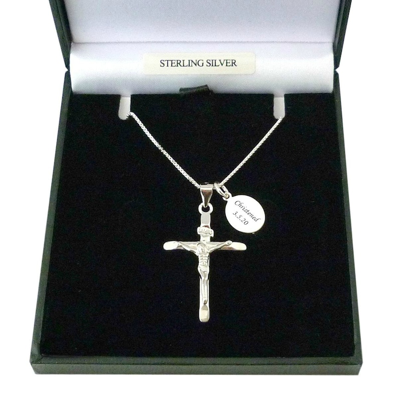 May include: A sterling silver crucifix pendant on a thin chain, presented in a black box with a label reading STERLING SILVER. An oval charm attached to the chain is engraved with the text Christened 3.5.20. The cross features a figure of Jesus.