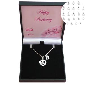 Könnte beinhalten: Eine silberne Halskette mit einem Herzanhänger und der Zahl 21, sowie einem Buchstaben "B" Charm, präsentiert in einer schwarzen Geschenkbox. Die Box hat ein rosa Innenfutter mit den Texten "Happy Birthday" und "With Love".