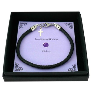 May include: A black braided leather bracelet with silver-toned metal end caps and a lobster clasp. A small silver-toned cross charm hangs from the clasp. The bracelet is displayed on a light purple card with the text To a Special Godson With Love.