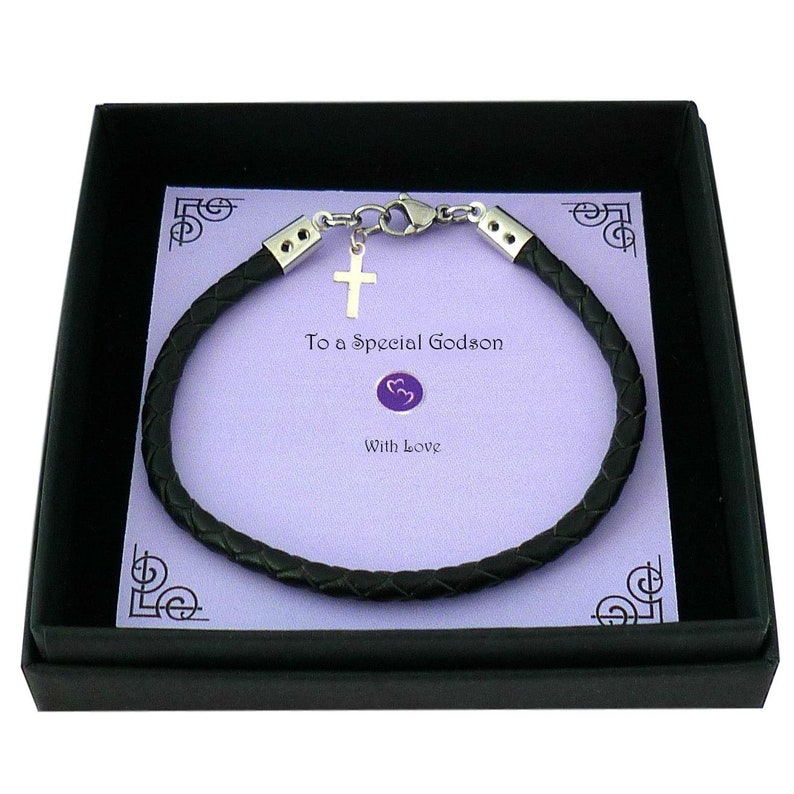 May include: A black braided leather bracelet with silver-toned metal end caps and a lobster clasp. A small silver-toned cross charm hangs from the clasp. The bracelet is displayed on a light purple card with the text To a Special Godson With Love.