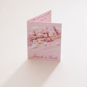 May include: A folded pink greeting card featuring a printed image of pink cherry blossoms and white pearls. The text on the front reads Jewels 4 Girls in a cursive font, and the interior text reads This gift was made especially for.