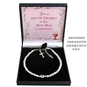 May include: A silver beaded bracelet with a cross charm and a letter charm inside a black gift box. The box has a pink interior with text that reads "For a Special Daughter on her First Holy Communion."