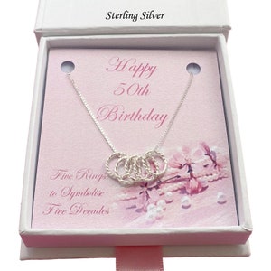 Könnte beinhalten: Eine silberne Halskette mit fünf ineinandergreifenden Ringen in einer weißen Geschenkbox. Die Box enthält eine rosa Karte mit der Aufschrift "Happy 50th Birthday" und "Five Rings to Symbolise Five Decades".