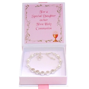 Puede incluir: Caja de regalo blanca con tapa rosa. La tapa tiene un borde floral y el texto "Para una Hija Especial en su Primera Comunión". La caja contiene una pulsera de plata con cuentas blancas, un dije de cruz de plata con un cristal transparente y cuentas de plata que deletrean el nombre "CLARA".
