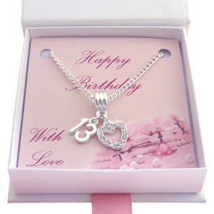 Peut inclure: Un collier argenté avec un pendentif en forme de chiffre 13 et un pendentif en forme de cœur orné de cristaux transparents. Le collier est présenté dans une boîte cadeau blanche avec une carte rose portant le texte Happy Birthday With Love.