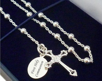 May include: A silver-toned rosary necklace featuring a series of small round beads on a chain. Attached to the chain is an ornate silver cross pendant and a small oval-shaped charm engraved with the text First Holy Communion in black lettering.