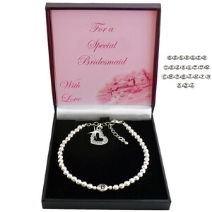 May include: A silver-toned beaded bracelet with a letter B charm and a heart-shaped crystal pendant inside a black gift box. The box lid features the text For a Special Bridesmaid With Love and a floral graphic. Includes a set of alphabet letter beads.