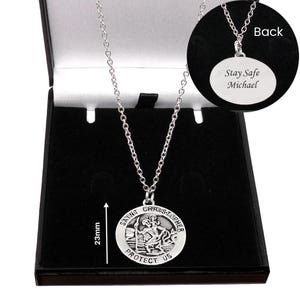May include: A silver-toned Saint Christopher pendant necklace on a chain. The circular pendant features a relief of Saint Christopher and the text 'SAINT CHRISTOPHER PROTECT US'. A 23mm measurement is indicated. The back is engraved 'Stay Safe Michael'.