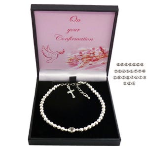 May include: A silver beaded bracelet with a cross charm and an initial letter charm, displayed in a black gift box. The box has a pink interior with the words "On your Confirmation" and a dove illustration.