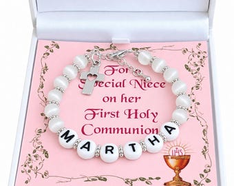 Può includere: Un braccialetto di perline bianche con un ciondolo a croce argentata con un cuore ritagliato e perline con lettere bianche che compongono il nome MARTHA. Il braccialetto è presentato su un cartoncino rosa con il testo For a Special Niece on her First Holy Communion.