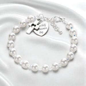 Könnte beinhalten: Ein Armband aus weißen Perlen mit einem silberfarbenen Kreuzanhänger und einem herzförmigen Anhänger, der mit dem Text First Holy Communion Day graviert ist. Das Armband verfügt über eine silberfarbene Metallkette mit einem Karabinerverschluss.