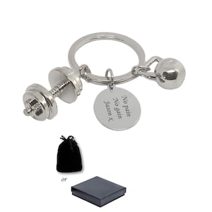 Könnte beinhalten: Ein silberfarbener Metallschlüsselanhänger mit einem Hantelanhänger, einem Kettlebell-Anhänger und einem runden Plättchen mit der Gravur No pain No gain Jason x. Das Set enthält einen schwarzen Samtbeutel oder eine schwarze Geschenkbox.