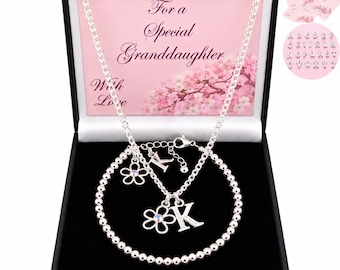 May include: A silver-toned chain necklace and a beaded bracelet set presented in a black gift box. The box insert features pink floral imagery and the text For a Special Granddaughter With Love. The jewellery includes flower and letter K charms.