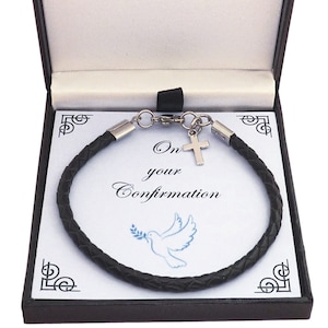 May include: A black braided bracelet with a silver-toned cross charm, presented in a black gift box. The insert card features the text On your Confirmation and a blue illustration of a dove holding an olive branch, with decorative corner accents.