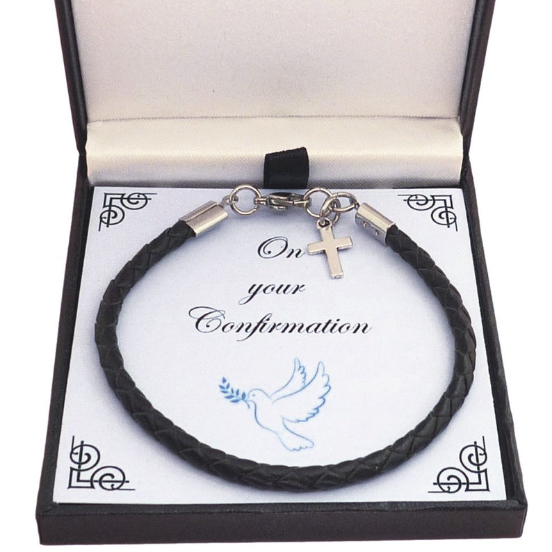 May include: A black braided bracelet with a silver-toned cross charm, presented in a black gift box. The insert card features the text On your Confirmation and a blue illustration of a dove holding an olive branch, with decorative corner accents.