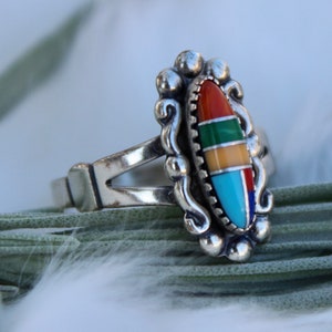 May include: A silver ring with a multi-colored stone inlay. The stone is oval shaped and features red, orange, yellow, green, blue, and turquoise colors.