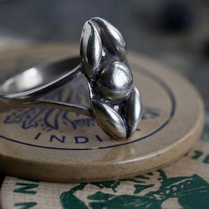 May include: A silver ring with a flower design. The ring is sitting on a brown and white surface with the word "INDI" visible.