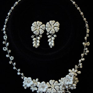 Op de afbeelding: Een zilveren ketting en oorbellenset met witte parel- en kristallen accenten. De ketting heeft een bloemmotief met delicate takken en bladeren. De oorbellen zijn van het druppeltype met een enkele bloem.