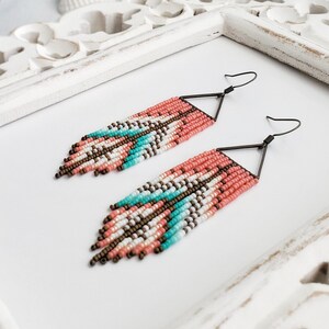 May include: A pair of long, beaded earrings with a geometric pattern in shades of coral, white, and turquoise. The earrings are made with small seed beads and have a triangle-shaped dangle at the bottom.